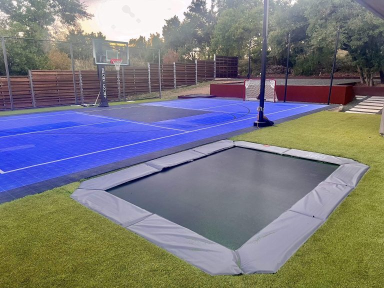 Outdoor blue sport court with basketball hoop, in-ground trampoline, and artificial turf in a residential backyard